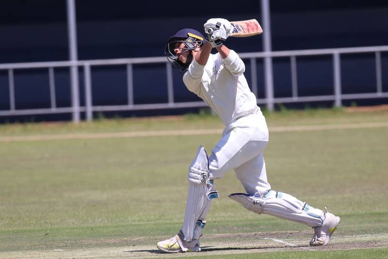 A batsman drives on a dusty maidan pitch, keeper at the stumps